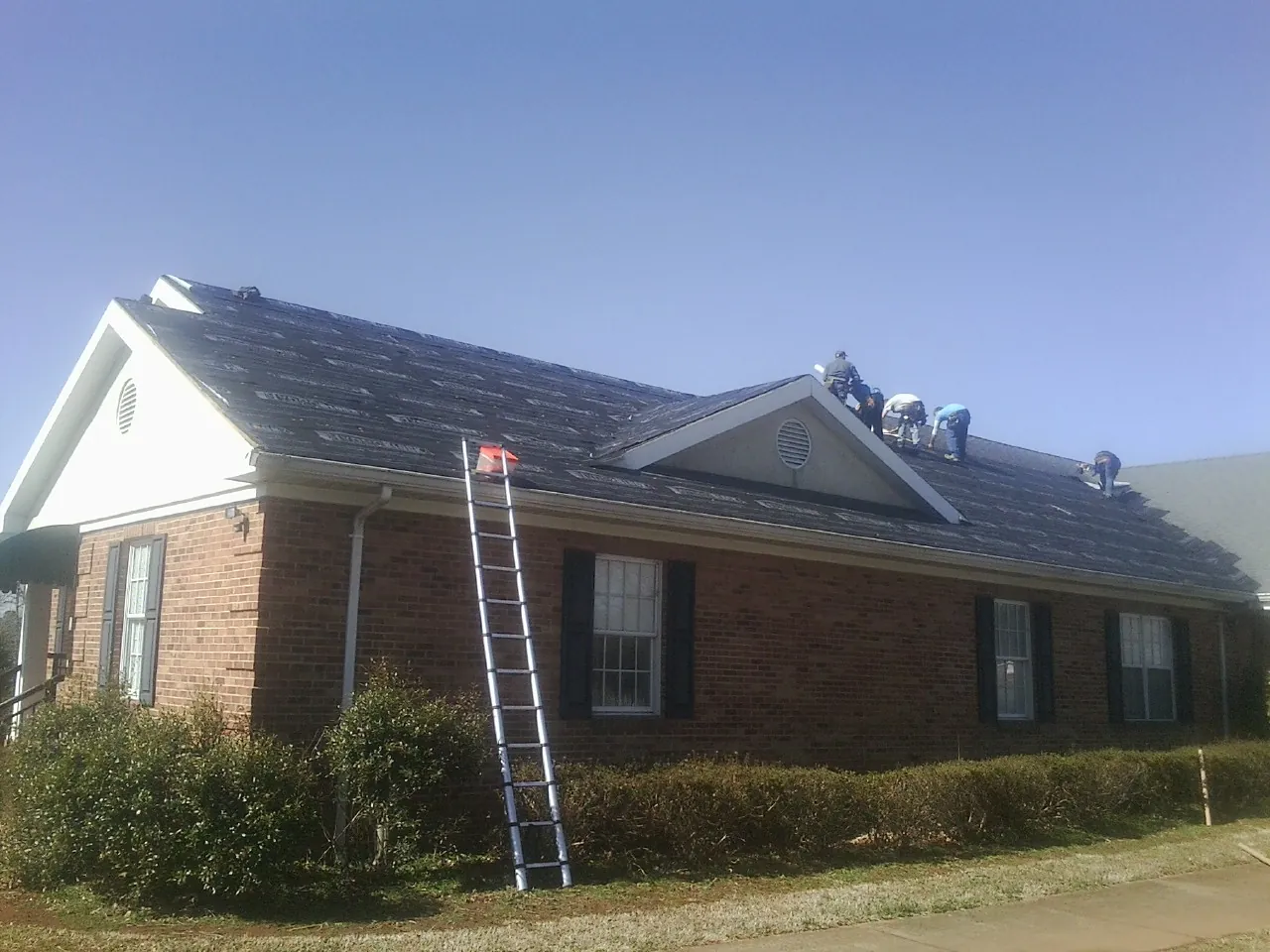 Crew tearing shingles off a brick ranch roof with a ladder set — Metal Roof Repair work by our crew in Locust Valley