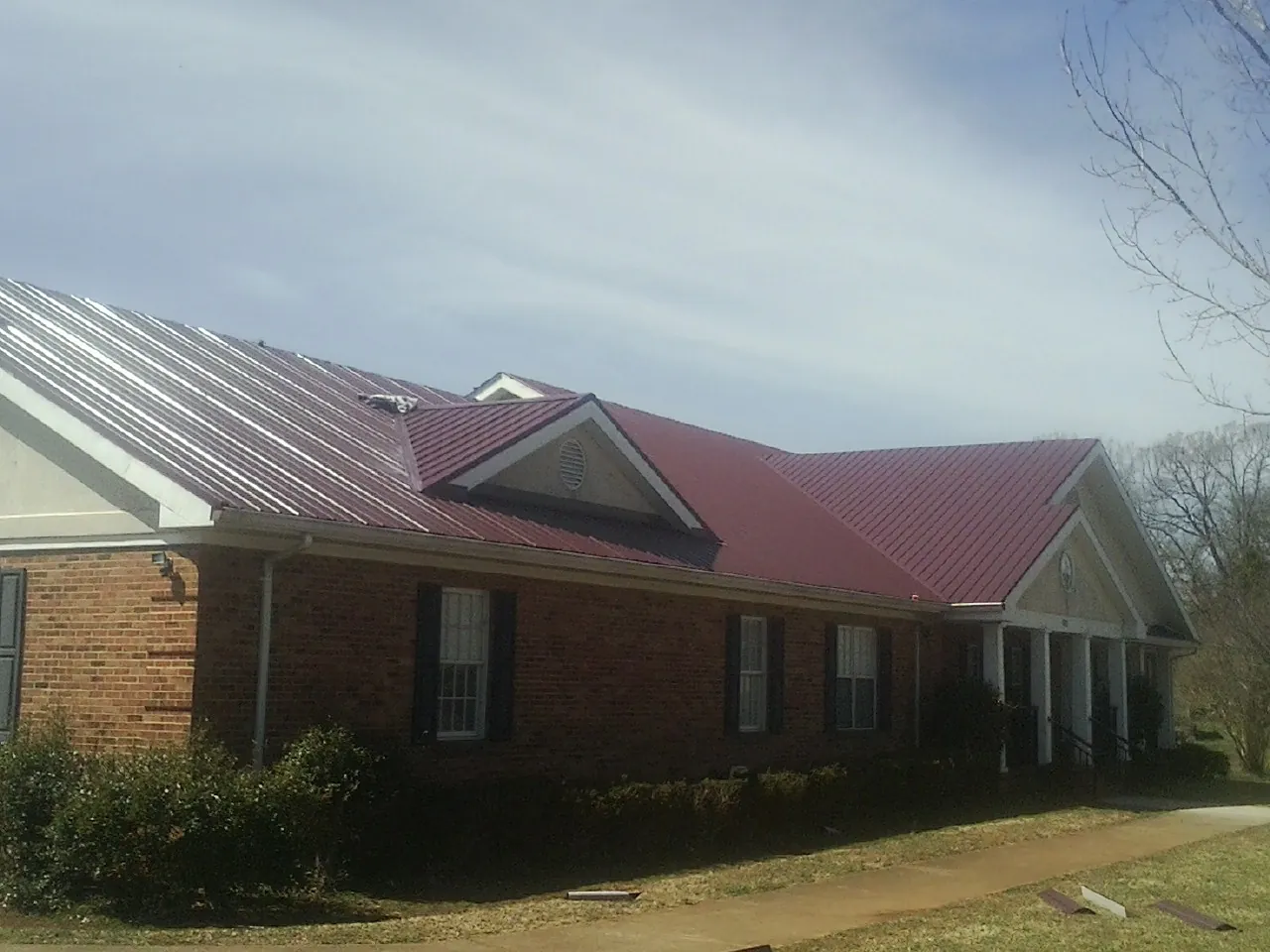 Red metal roof with gabled entries on a long brick building, from a Hail Damage Roof Repair job in Locust Valley