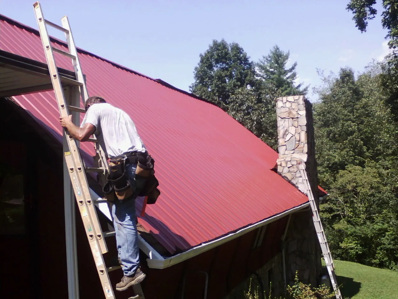 Roofer on a ladder beside a red metal roof and stone chimney — Soffit & Fascia Repair by Roofing Brothers in Locust Valley