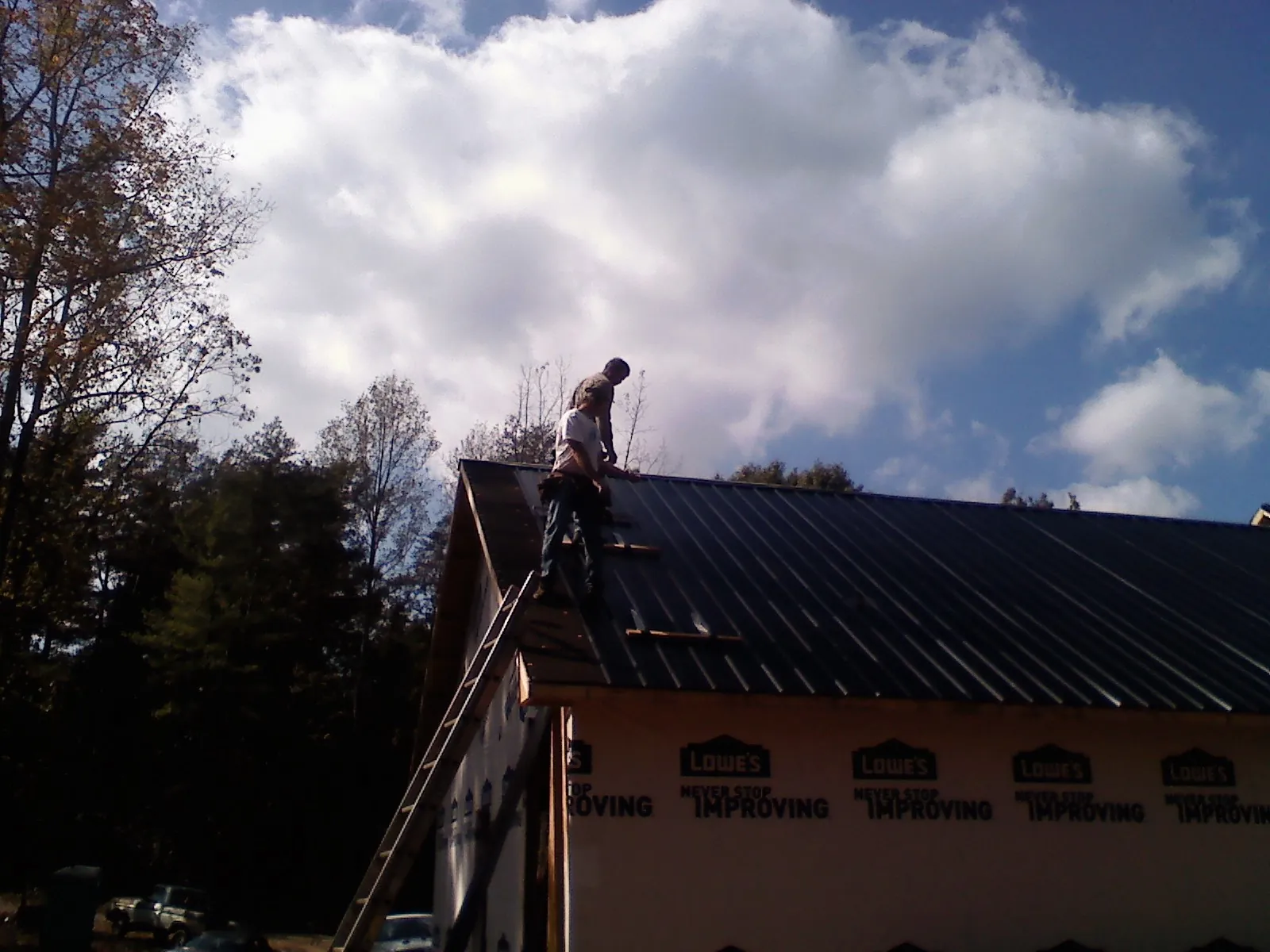 Roofer fastening dark metal panels high against a cloudy sky, from a Roof Inspection job in Locust Valley